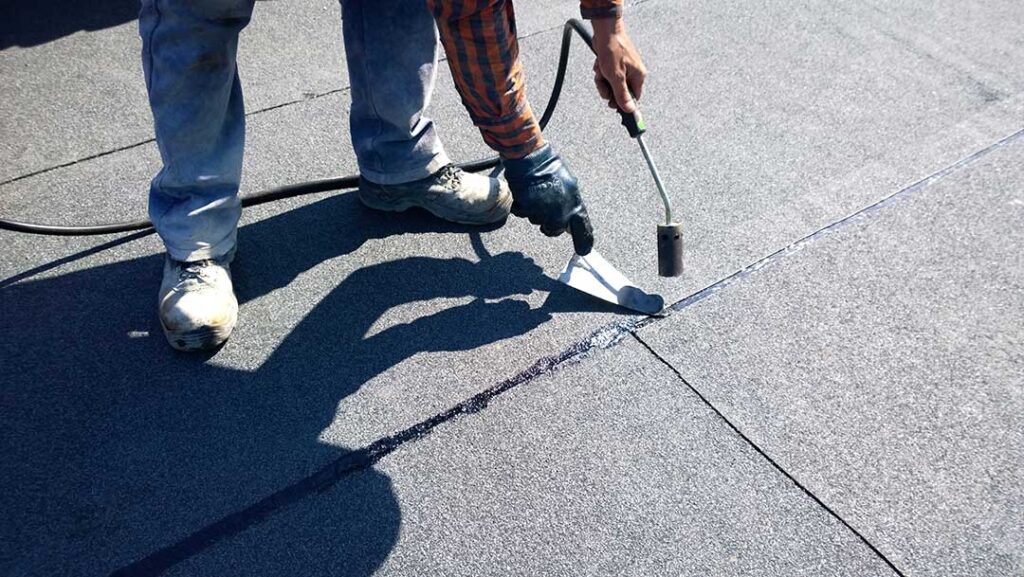 A homeowner using a level on a shingle roof to check the slope angle on a sunny day.
