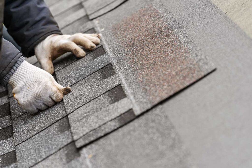 A homeowner in casual clothes inspects their roof for storm damage from a backyard, with missing shingles visible.
