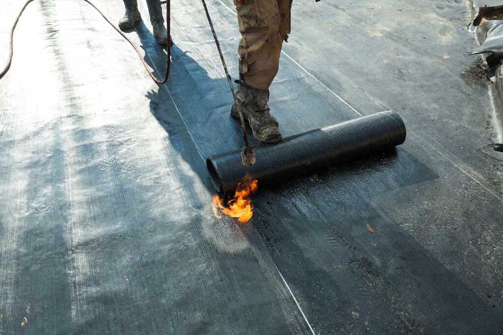 Roofing workers inspect exposed roof decking beside stacked shingles and underlayment materials.